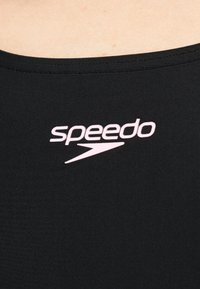Μαύρο μαγιό από ύφασμα με λευκό λογότυπο Speedo τυπωμένο στο στήθος, ορατός λαιμός και άνω μέρος του στήθους.