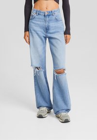 Donna che indossa jeans a vita alta di colore azzurro chiaro, larghi e con ampie strappi sulle ginocchia, abbinati a sneakers grigie e a un top nero a maniche lunghe cropped.