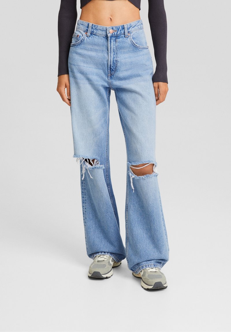 Donna che indossa jeans a vita alta di colore azzurro chiaro, larghi e con ampie strappi sulle ginocchia, abbinati a sneakers grigie e a un top nero a maniche lunghe cropped.