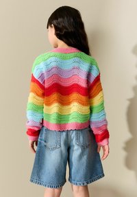 Pull tricoté multicolore avec des rayures ondulées vertes, roses, orange et bleues, associé à un short en denim bleu clair.