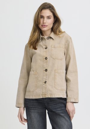 Žena v béžovom zapínacom saku s tromi prednými vreckami a tmavých džínsoch, stojaca pred jednoduchým svetlým pozadím.