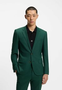 Grüne Anzugjacke mit taillierter Passform, ausgestattet mit goldenen Knöpfen, zwei Fronttaschen und einem schwarzen strukturierten Poloshirt darunter.