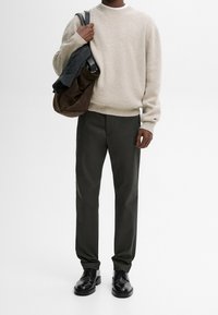 Pull en laine beige, pantalon vert foncé, chaussures en cuir noir, tenant un sac marron avec un pull gris posé dessus. Fond neutre.