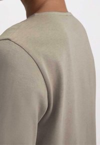 Närbild av en persons övre rygg och axel som bär en beige långärmad skjorta med synliga sömmdetaljer.