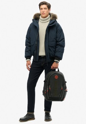 Veste matelassée marine avec capuche en fourrure, col roulé crème, jeans foncés, chaussures noires et un sac à dos noir avec des accents orange et un logo.