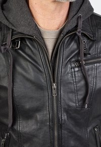 Zwarte leren jas met een ritssluiting aan de voorkant, een grijze capuchonvoering, zijzakken en gestructureerd leer met zichtbare stikseldetails.