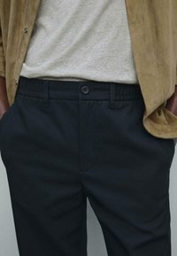 Dark navy black trousers featuring an elastic waistband, single button closure, and side pockets, paired with a light gray shirt and tan jacket.