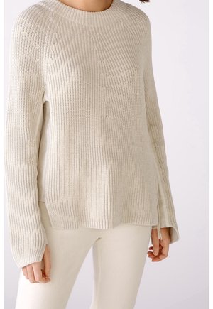 Kvinde iført en cremefarvet strikket sweater med sideslidser og matchende cremefarvede bukser mod en ensfarvet baggrund.