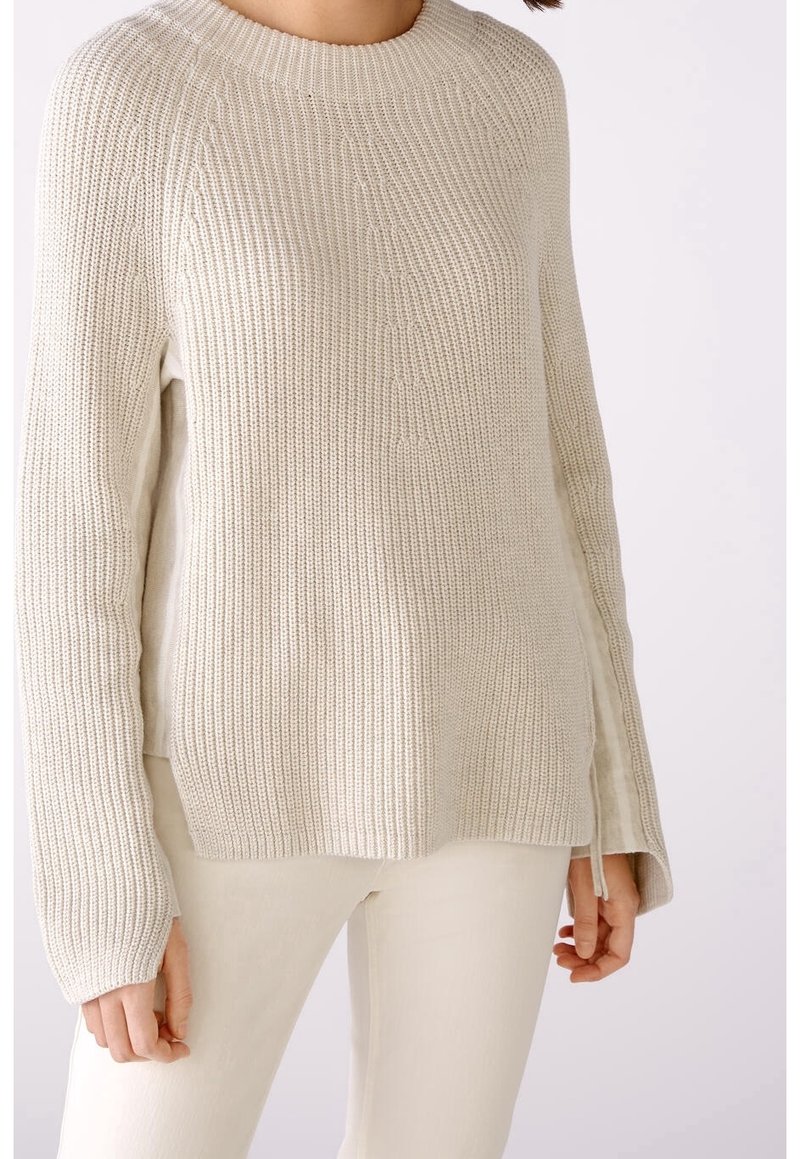 Femme portant un pull en maille crème avec des fentes sur les côtés et un pantalon crème assorti sur un fond uni.