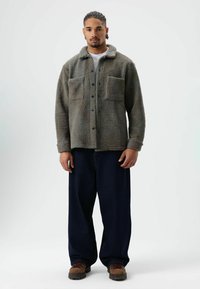 Fleece shirt jacket in muted green, featuring a collar, two large chest pockets, front buttons, and a textured surface. Paired with loose blue pants.