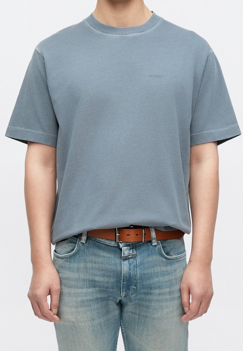 Personne portant un t-shirt à manches courtes bleu clair rentré dans un jean bleu délavé avec une ceinture marron.