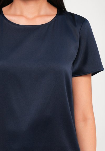 Woman wearing a short-sleeve navy blue satin blouse with a round neckline, visible from shoulders to mid-torso.
