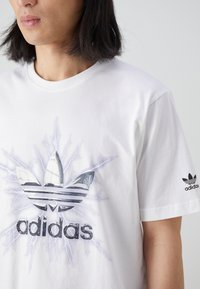 Bílý bavlněný tričko s velkým, stylizovaným černým a šedým logem Adidas obklopeným grafikou blesku; krátké rukávy a kulatý výstřih.