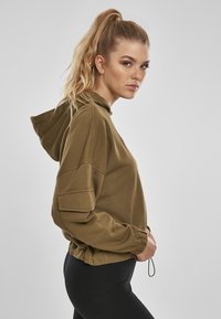 Sudadera verde oliva hecha de una tela suave, con capucha ajustable, hombros caídos y puños acanalados. Diseño elegante con un corte recortado.