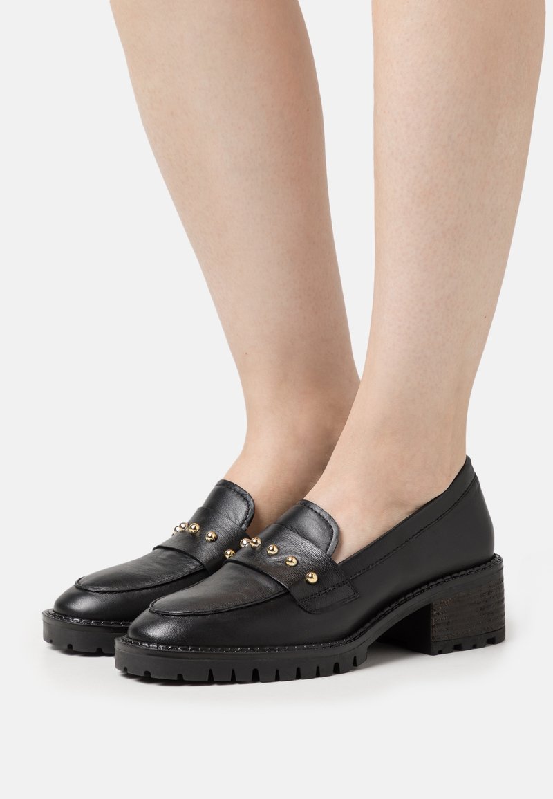 Mocassins noirs en cuir à talon bas avec des clous dorés portés aux pieds, dotés d'une semelle épaisse à crampons et d'un design à bout arrondi.