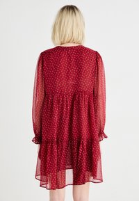 Rochie roșie semi-transparentă cu imprimeu floral, mâneci bufante, decolteu rotund, fustă în straturi și manșete cu volane. Material ușor, texturat.