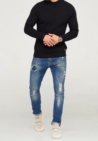 Pull en tricot noir, jean bleu déchiré avec des déchirures, et baskets claires avec des bretelles. La tenue présente un design décontracté et ajusté.