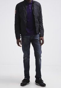 Svart bomberjacka med kviltade detaljer på axlarna och bröstfickor, bar över en lila polo, parat med blå jeans och svarta sneakers.