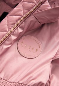 Innenansicht einer rosa gesteppte Jacke mit einem runden Stoffetikett und geprägter goldener Schrift, das "BAKER by Ted Baker, Vereinigtes Königreich, London" anzeigt.