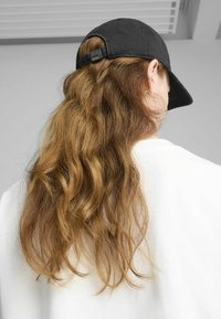 Black baseball cap with a curved visor, worn backward, highlighting wavy, long brown hair and a white sweater in a neutral background.