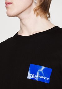 Melnas kokvilnas T-krekls ar apaļu kakla izgriezumu. Iezīmēts ar spīdīgu zilu taisnstūra birku ar uzrakstu "KARL LAGERFELD JEANS" baltā krāsā.