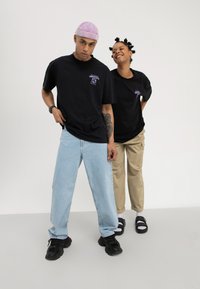 Twee jonge volwassenen die zwarte grafische T-shirts dragen; de een in lichtblauwe jeans en een paarse pet, de ander in beige broek met sandalen en sokken, poserend in casual houding.