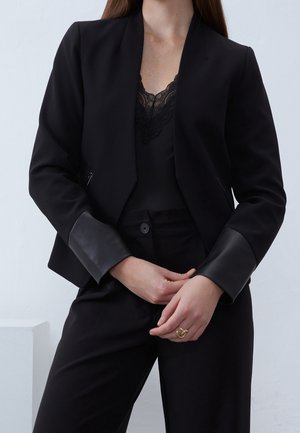 Kvinde iført sort blazer med lædermanchetter, sort blondekantet top og sorte bukser, hænderne foldet ved taljen, neutralt baggrund.