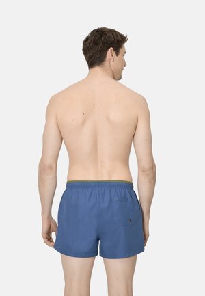 Hombre con la espalda desnuda vistiendo pantalones cortos de baño azules, de pie de espaldas contra un fondo claro y liso.