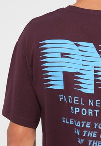 Bordeaux bombažna majica s sešnjo v modri barvi, ki vsebuje izstopajoč grafični dizajn z linijskimi detajli in besedilom "PADDEL NEW SPORT."