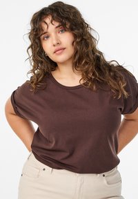 Braunes Baumwoll-T-Shirt mit lockerem Schnitt und kurzen Ärmeln, kombiniert mit hellfarbigen Hosen. Der Stoff wirkt weich mit minimaler Textur.