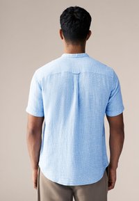 Hele sinine lühikeste varrukatega särk, mis on valmistatud tekstuurse kangaga, omades ümarat ääreosa ja tagaosa detailis keerukat lukut.