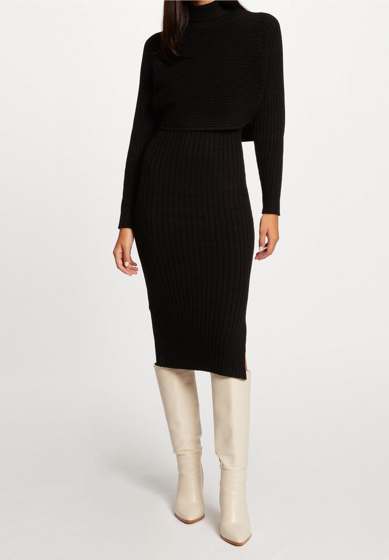 Femme portant une robe midi en maille côtelée noire et des bottes hautes crème, debout devant un fond blanc.