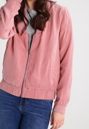 Kobieta nosząca różową kurtkę sztruksową na zamek błyskawiczny na szarym t-shircie i niebieskich dżinsach, z częściowo widocznymi długimi ciemnymi włosami.