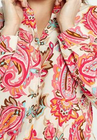 Floral blouse featuring a cream background with vibrant pink, blue, and brown paisley patterns. Includes button closures and a mandarin collar.