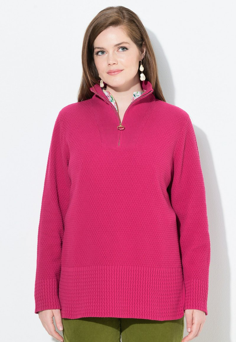 Donna con lunghi capelli castani che indossa orecchini a goccia di perle, un maglione rosa brillante con texture e mezza cerniera, e pantaloni verde oliva.
