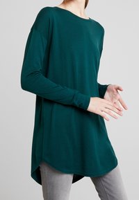 Türkisfarbenes Langarmshirt mit rundem Ausschnitt und abgerundetem Saum aus weichem Stoff; getragen über grauen Jeans, mit lockerem Schnitt und Seitenschlitzen.