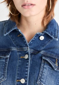 Veste en denim bleu avec un col, boutons-pression argentés, poches avant et détails de couture jaune visibles. Texture lisse.