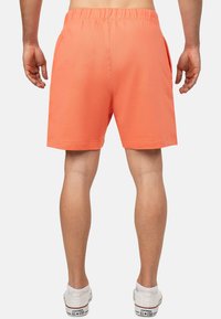 Oranje sportshorts met een elastische tailleband, zijzakken en een gladde textuur. Draagend met witte sneakers, gezien vanuit de achterkant.