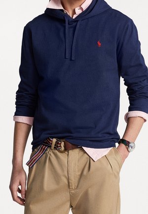 Hombre con sudadera con capucha azul marino sobre camisa rosa claro de botones y pantalones beige con cinturón de tela a rayas y reloj de pulsera, manos en los bolsillos.