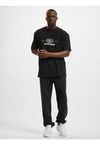 Svart oversized t-skjorte med "SPACE BUDS" grafikk, kombinert med svarte joggebukser. Hvite sneakers fullfører antrekket. Glatt, uformelt stoff.