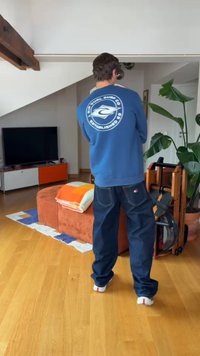 Homme portant un sweat-shirt bleu de Surf Company et un jean se tenant dans un salon avec un ottoman, une télévision, une plante et un rameur.