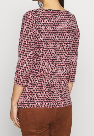 Femme portant un haut à manches longues multicolore à motifs et un pantalon marron, vue de dos sur un fond uni.