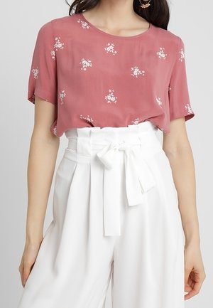 Kobieta w różowej bluzce w kwiaty z krótkim rękawem, wpuszczonej w białe, plisowane spodnie z wysokim stanem i wiązanym materiałowym paskiem.