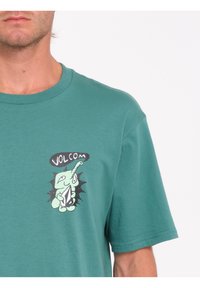 Camiseta de algodón verde con un gráfico de elefante de dibujos animados en blanco y negro con el texto "VOLCOM" encima, cuello redondo y mangas cortas.