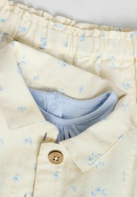 Cămașă galben deschis cu un model floral albastru, având un sistem de închidere cu nasturi și un guler interior albastru. Materialul are un aspect texturat.
