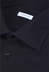 Gros plan sur le col texturé foncé d'une chemise avec une petite étiquette blanche portant la mention "seidensticker" à l'intérieur de l'encolure.