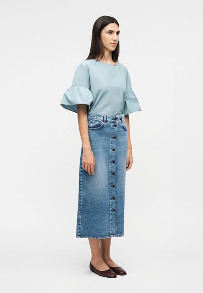 Γυναίκα που φοράει μπλούζα με φουσκωτά μανίκια σε ανοιχτό γαλάζιο, μεσαίο μήκος φούστα από τζιν με κουμπιά μπροστά και σκούρα επίπεδα παπούτσια, στέκεται μπροστά σε απλό λευκό φόντο.