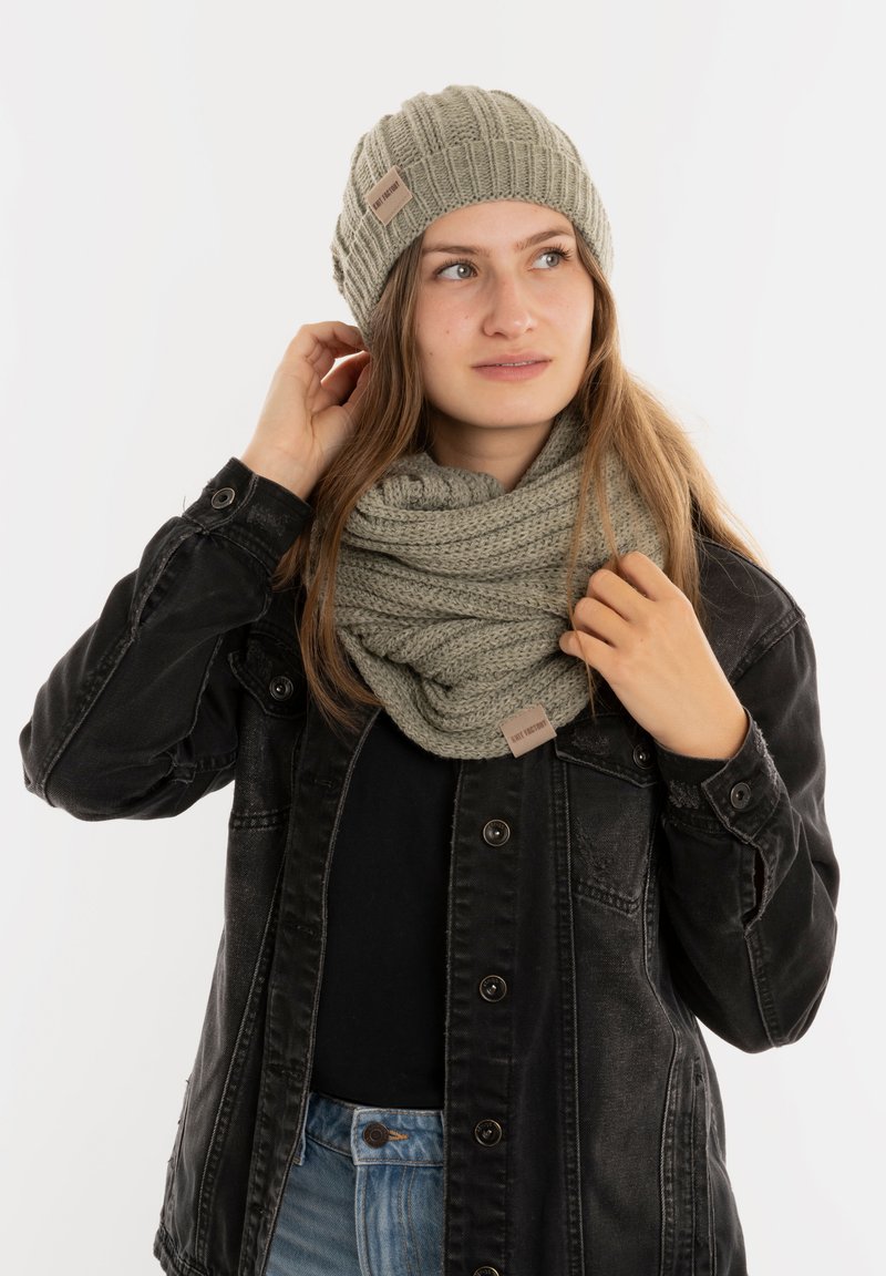 Gehaakte olijfgroene beanie met ribbels en label, gecombineerd met een textuurgehaakte infinity-sjaal. Model draagt een zwarte jas en blauwe jeans.