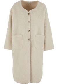 Bēša mākslīgās kažokādas mētelis ar apaļu kakla izgriezumu, priekšpusē aizdare ar pogām, īsām piedurknēm un divām kabatām. Mīksts faktūra un vaļīgs dizains.