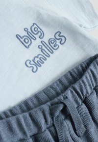 Tissu bleu clair avec texte brodé "big Smiles" accompagné d'un pantalon en maille côtelée bleu foncé avec ceinture à cordon.